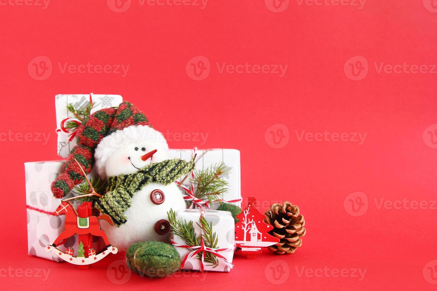 Christmas still life with snowman and gift boxes wrapped of white-gray paper with red-white ribbons on a red background, decorated of fir branches, pine cone and red wooden decorations. photo