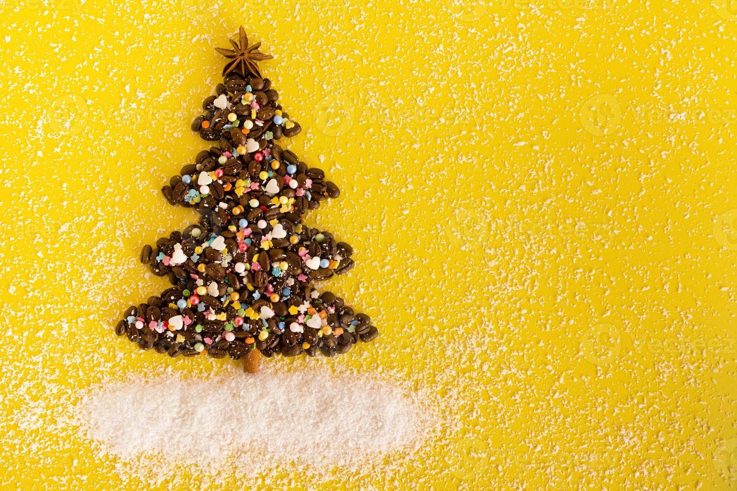 árbol de navidad hecho de granos de café y estrella de anís decorada, aspersión culinaria multicolor y chips de coco sobre un fondo amarillo, vista superior. foto