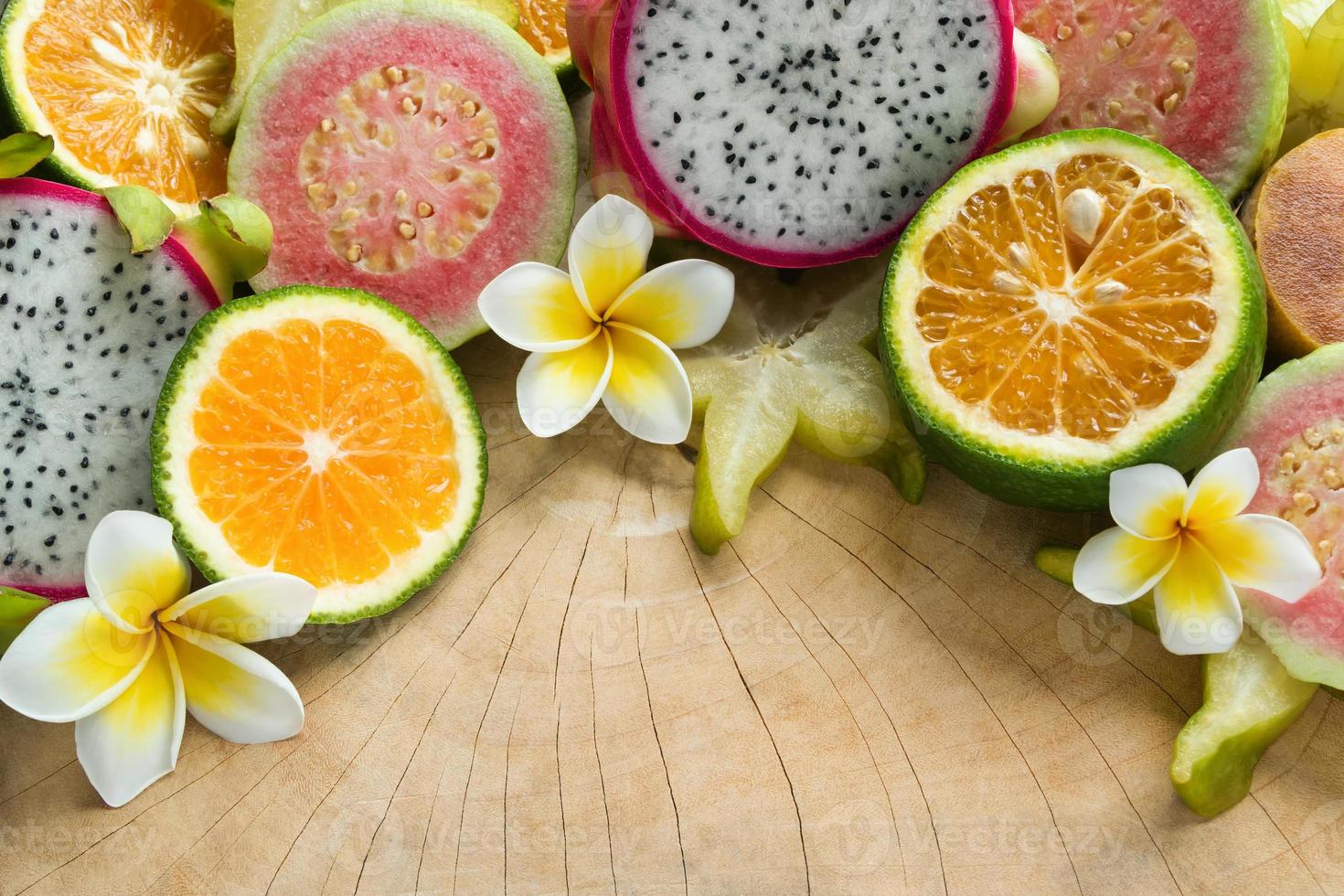 frutas tropicales de colores brillantes - mandarina, guayaba, fruta de dragón, fruta de estrella, sapodilla con flores de plumeria en el fondo de madera. foto