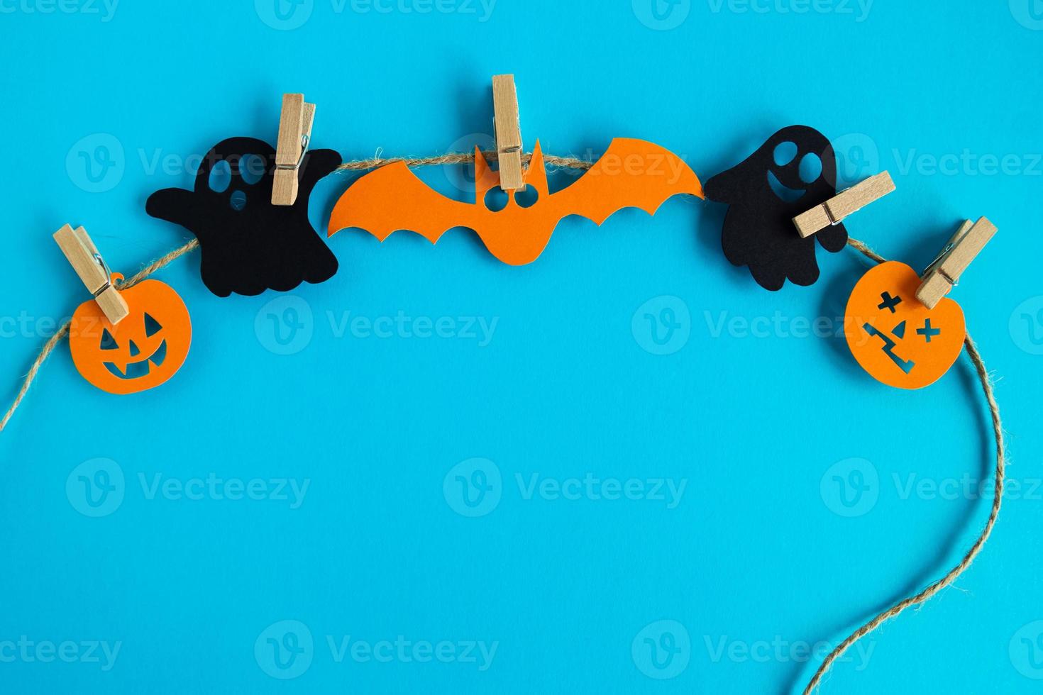 Holiday decorations for Halloween. Black paper ghosts and orange paper pumpkins and bat hanging on a rope on a blue background with copy space. photo