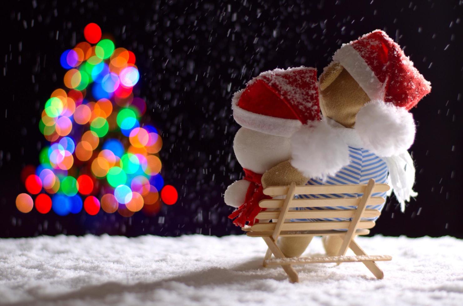 Brown and white teddy bear wearing santa hat sitting on wooden bench when snowing in winter looking at colorful bokeh lights of Christmas tree. photo