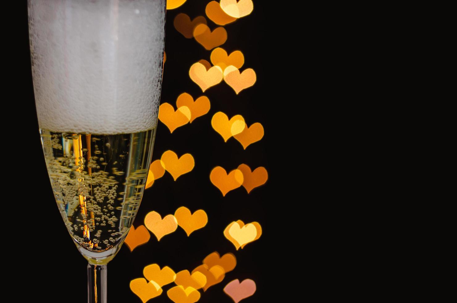 Blurred focus of a glass of sparkling wine with white bubble and love shape golden bokeh lights on dark background. photo