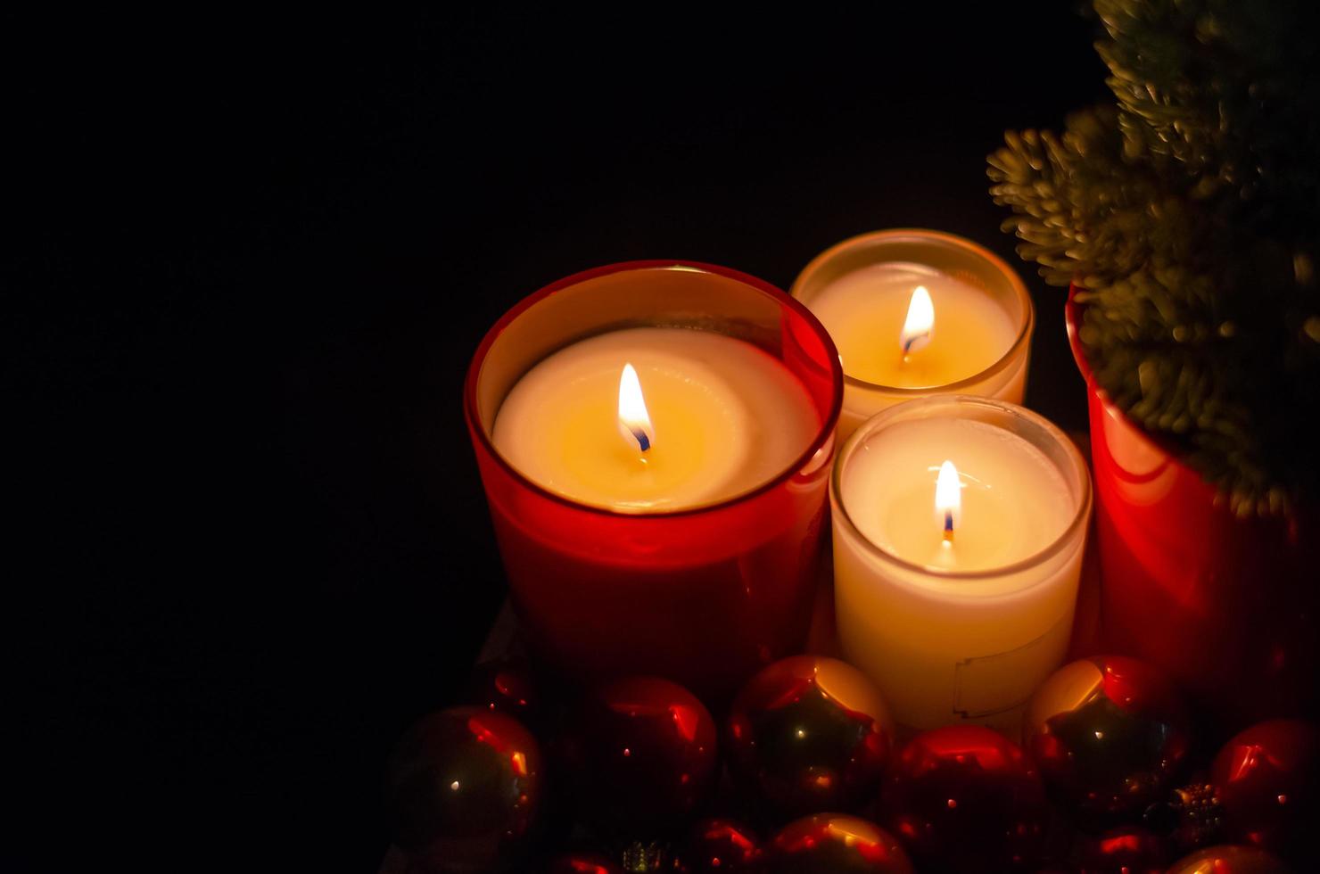 Selective focus on flame burning of red glass candle with Christmas tree and bauble ornaments in dark background. photo