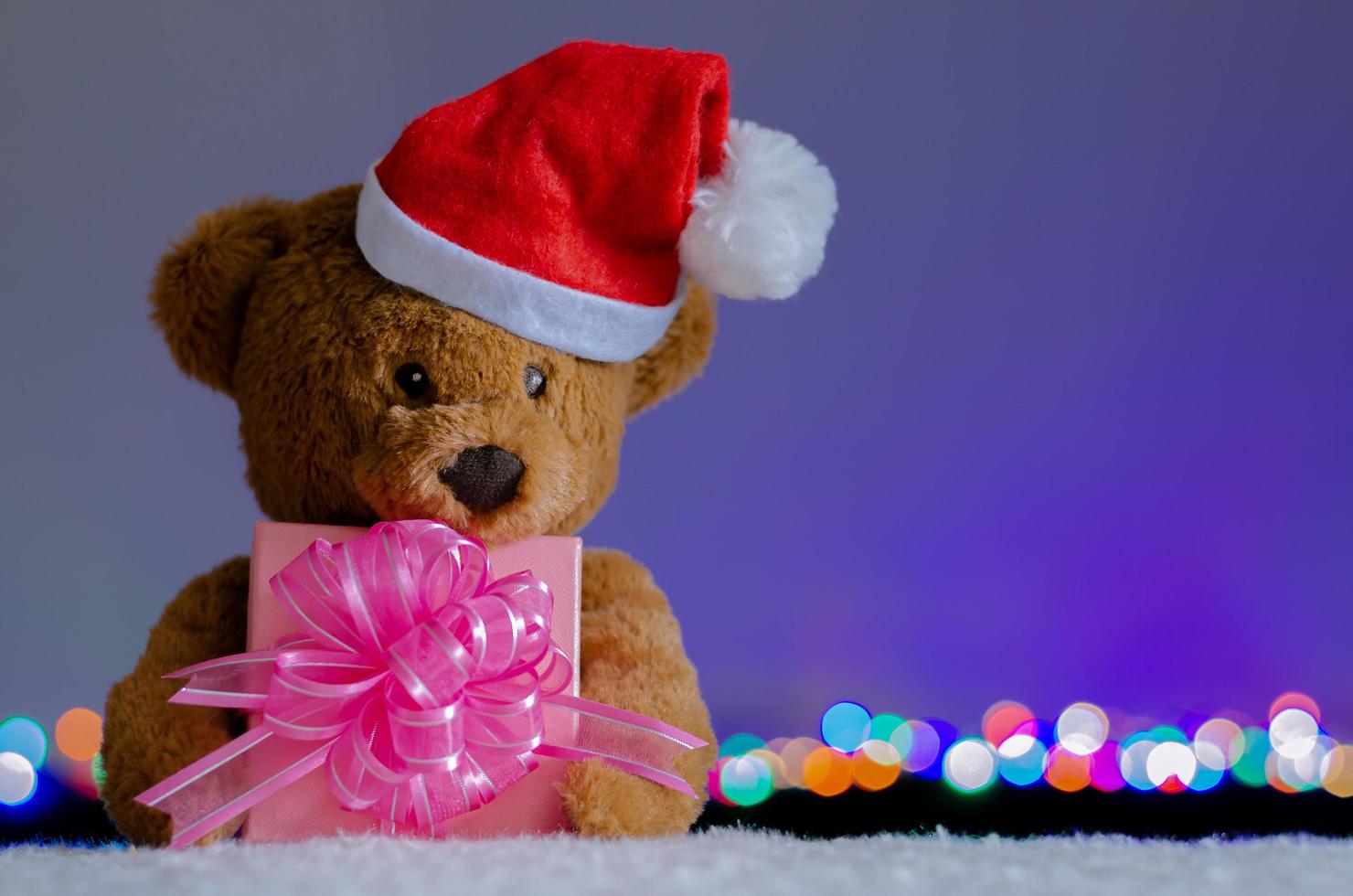 Brown teddy bear wearing santa claus hat holding partial focus of Christmas gift box and bokeh lights background. photo