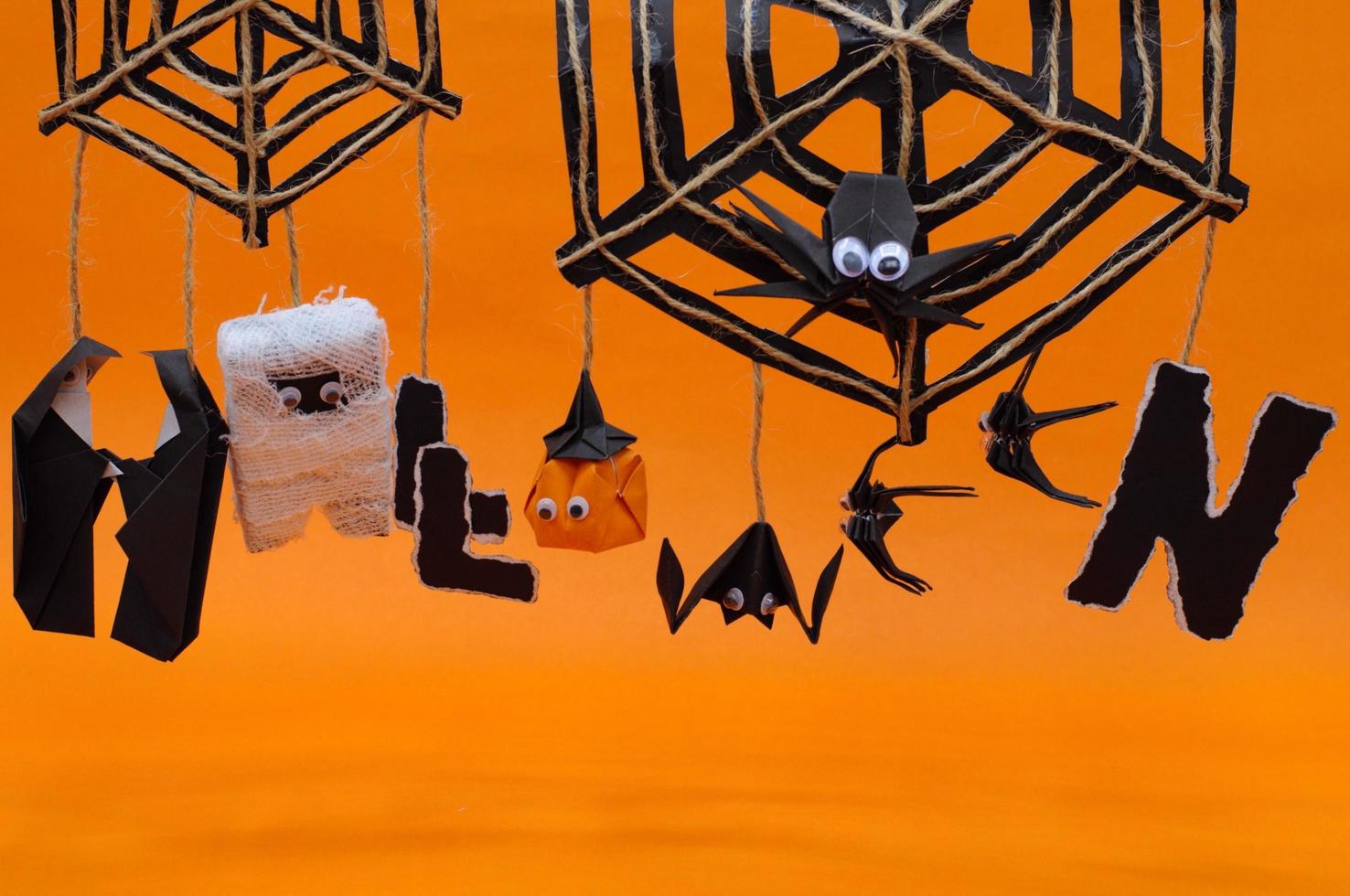 The origami Halloween background of pumpkin head jack o lantern mummy bat and nun hanging on spider cobweb set to the word HALLOWEEN isolated on orange. photo