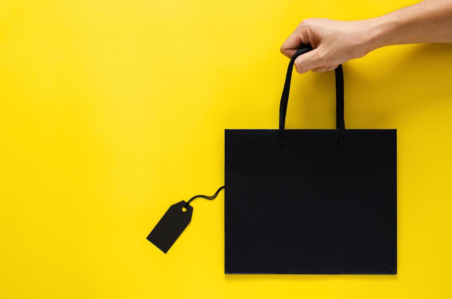 Hand holding a black shopping paper bag with blank black price tags on yellow background for Black Friday shopping sale concept. photo