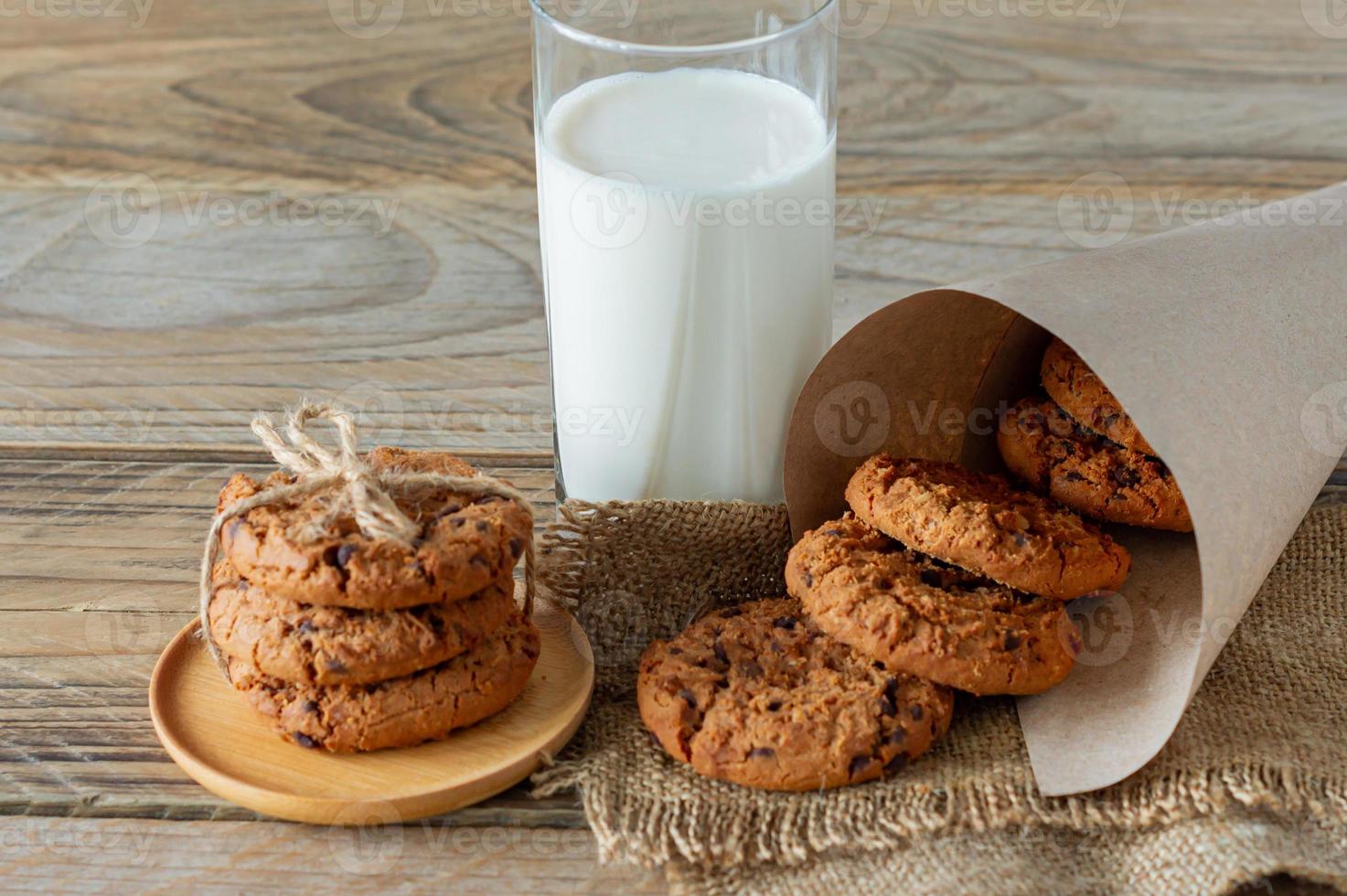 A stack of chocolate chips cookies on baking paper, with a glass of milk on a light wooden background photo