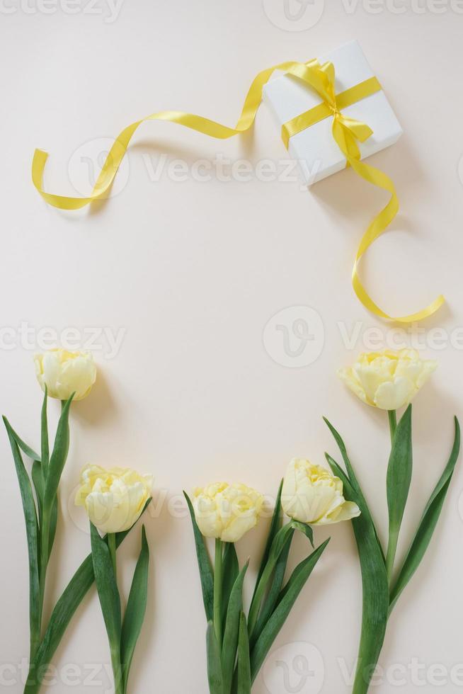 Gift box wrapped in white paper with a yellow ribbon and flowers of yellow tulips on a light table. Flat lay. Copy space for text. photo