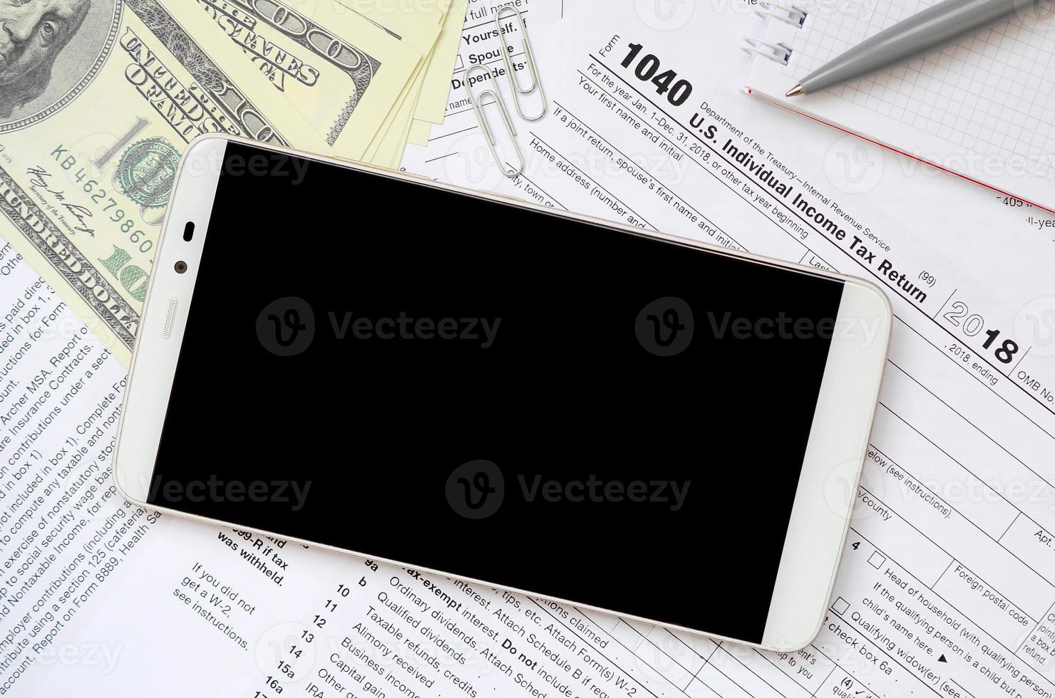 Composition of items lying on the 1040 tax form. Dollar bills, pen, smartphone, paper clip and notepad. Clean black screen smartphone for text placement photo