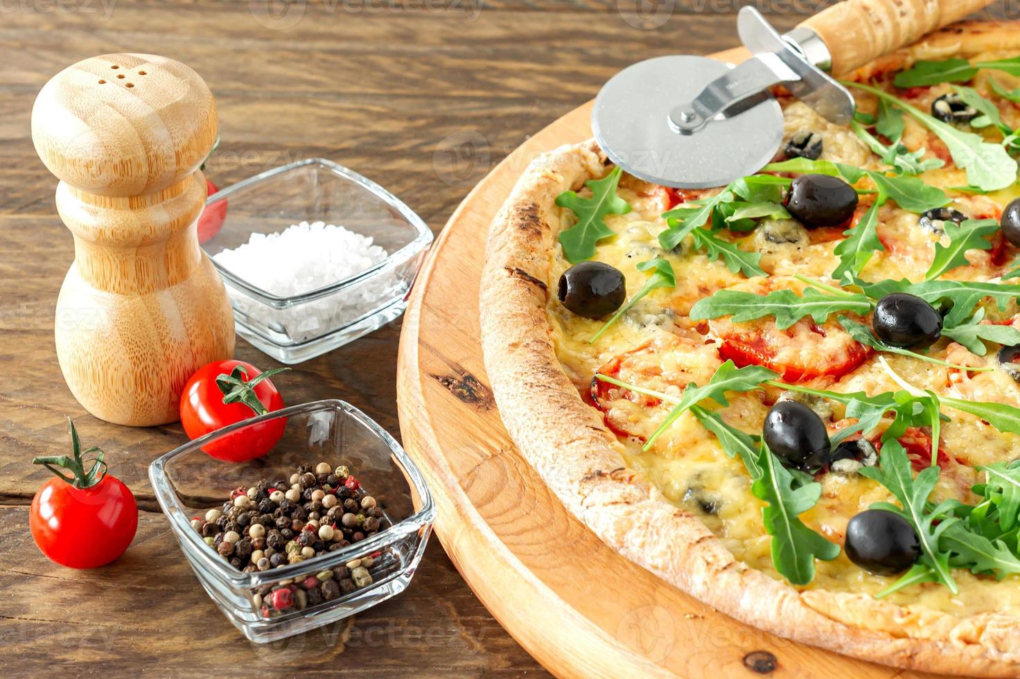 cooked hot pizza pie with pepperoni, tomatoes, black olives, arugula and mozzarella on wooden board. photo