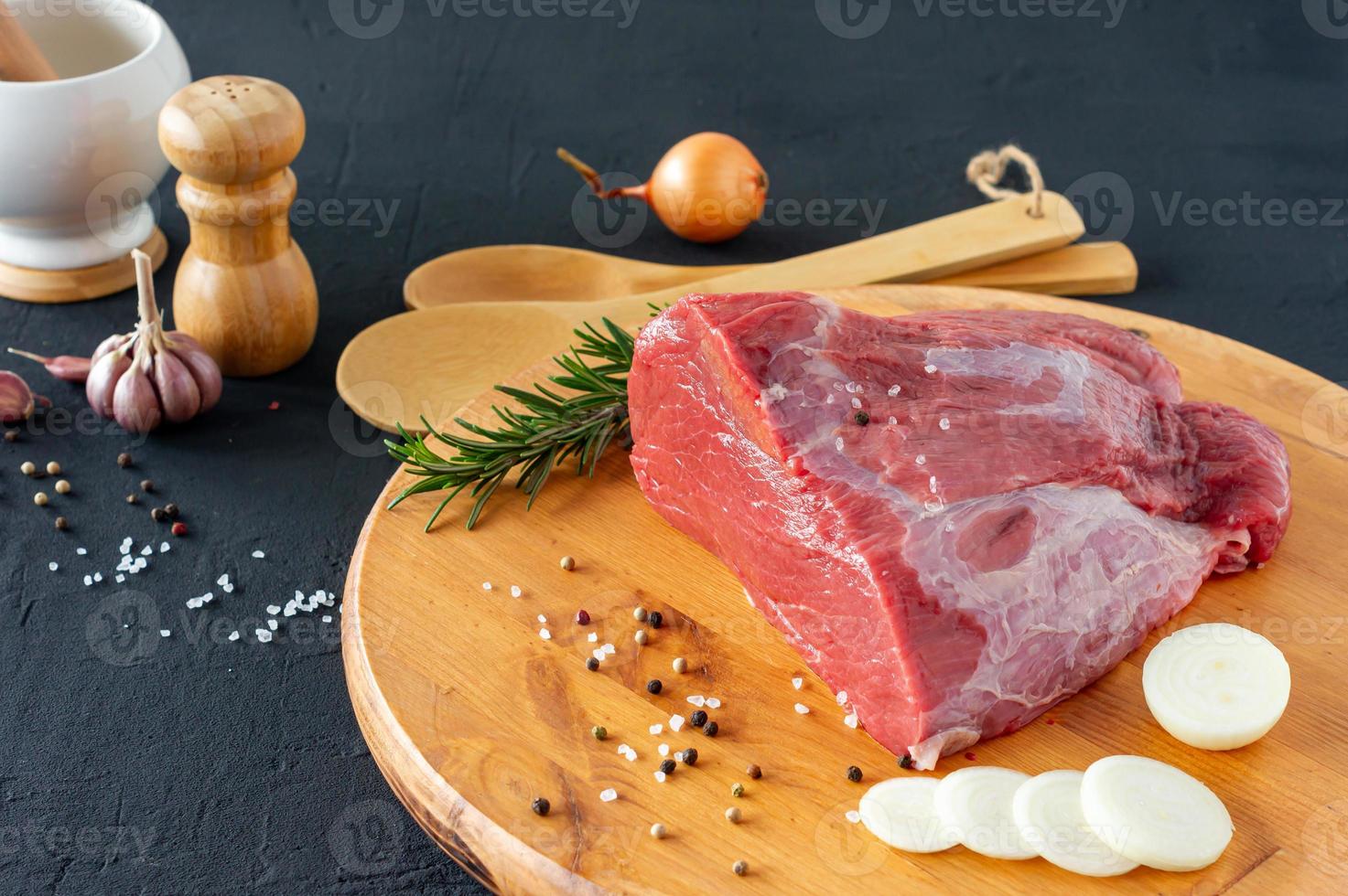 raw meat beef fillet on wooden cutting board, rosemary twigs, different peppers, salt and onion on dark background. Fresh beef piece for steak or grilled barbecue photo