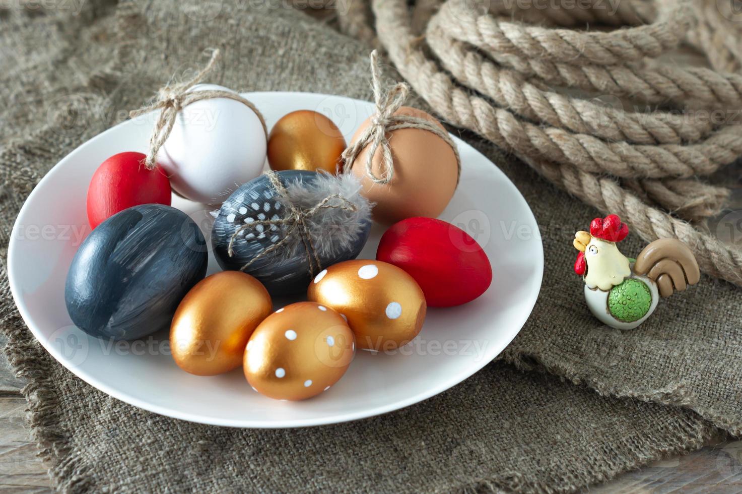 huevos de pascua de colores modernos, lienzo y cuerda y plumas en un plato sobre una mesa de madera. preparación para las vacaciones de semana santa. composición festiva. foto