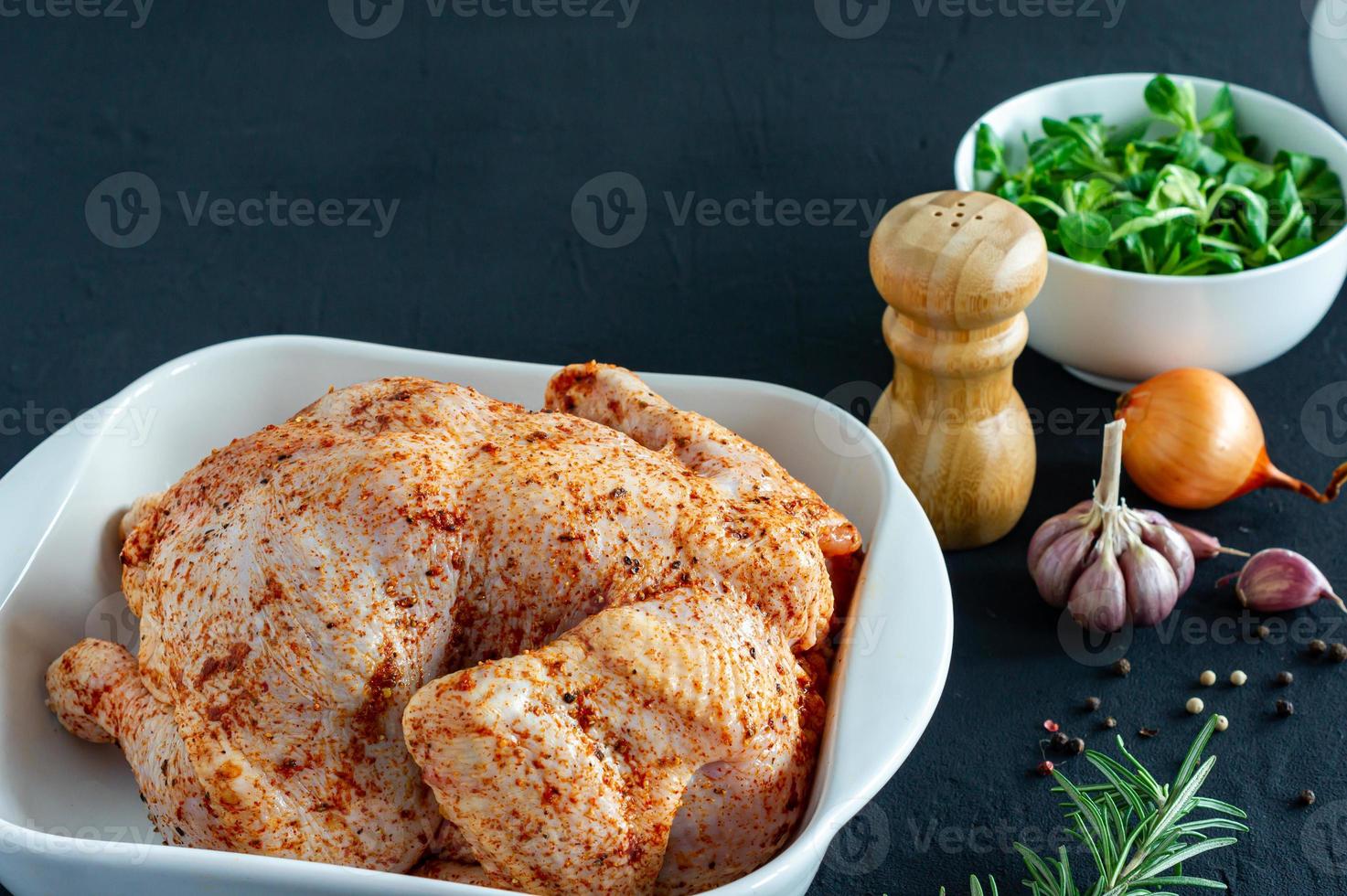 Whole raw chicken with herbs and spices ingredients in baking ceramic dish on dark background ready to cook. photo