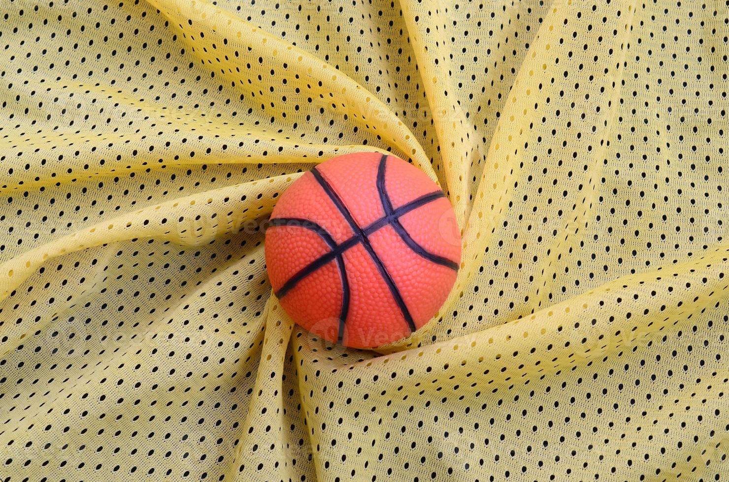 Small orange rubber basketball lies on a yellow sport jersey clothing fabric texture and background with many folds photo