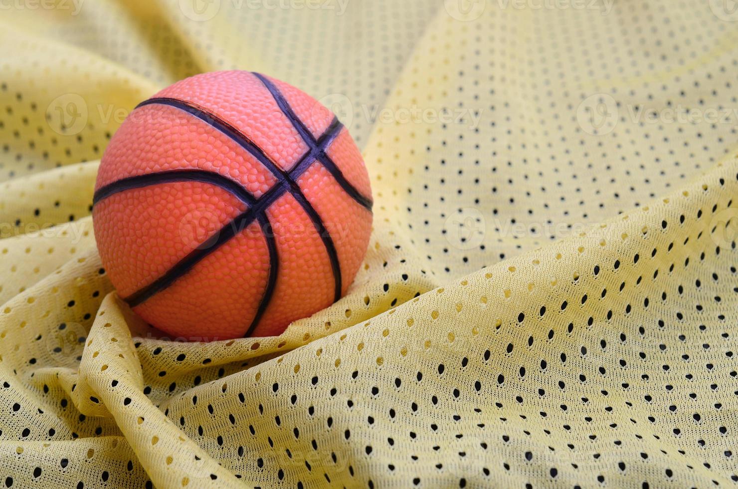 el pequeño baloncesto de goma naranja se encuentra sobre una textura de tela de jersey deportivo amarillo y un fondo con muchos pliegues foto