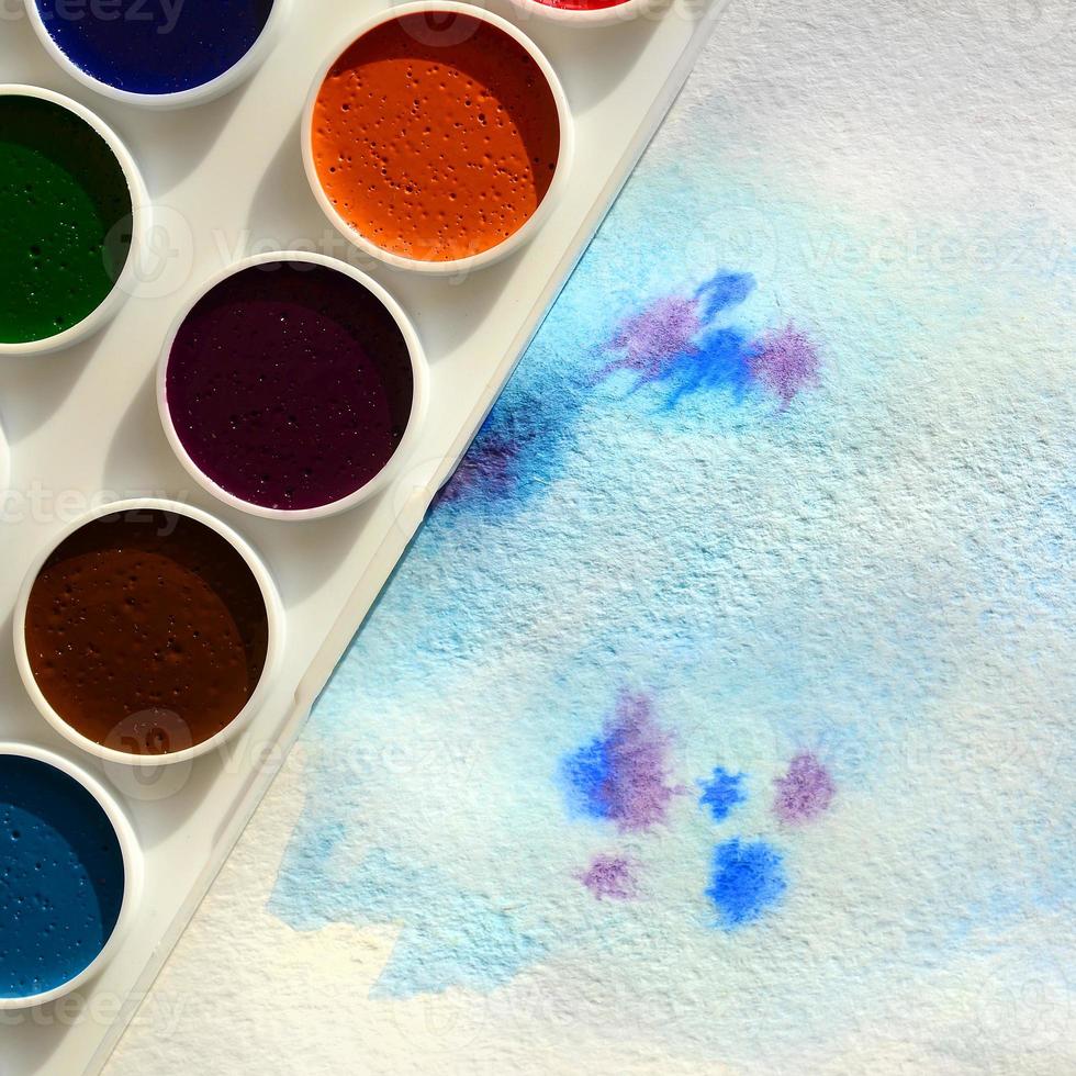 A new set of watercolors lies on a sheet of paper, which shows an abstract watercolor drawing in the form of blue strokes. The concept of amateur painting among teenagers photo