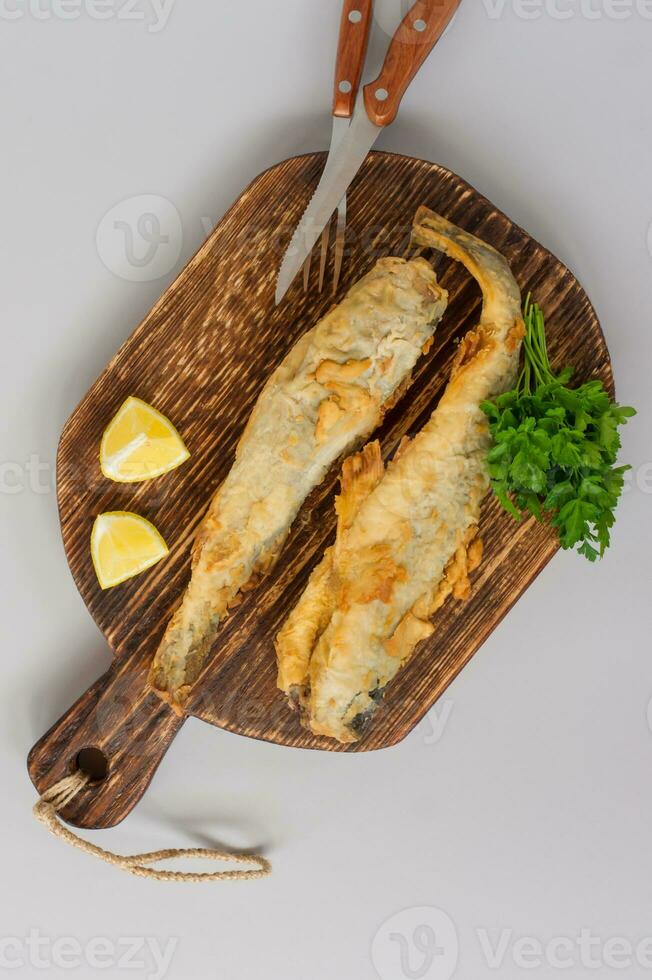 vista superior de navaga de pescado frito servido en tabla de cortar de madera con limón, verduras y verduras sobre fondo gris foto