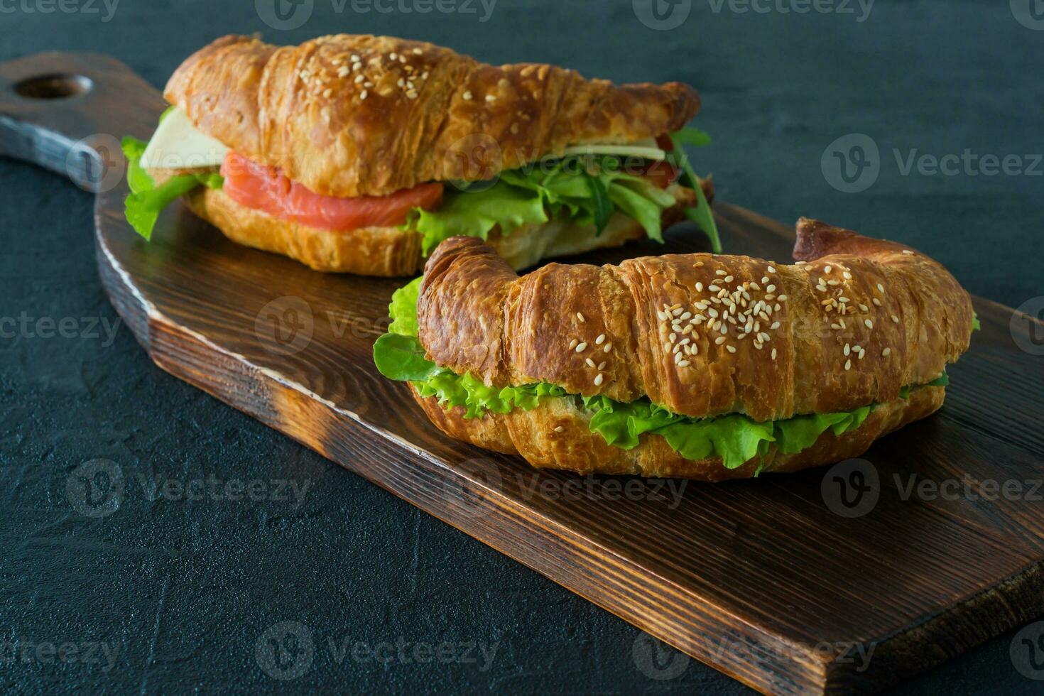 Croissant sandwiches with salted salmon on a desk, served with fresh salad leaves, arugula and vegetables over black background photo