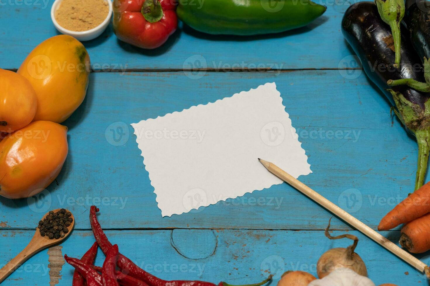 Vegetables are laid out around a sheet of paper and a pencil. Empty space for text. Vegetables, empty blank for recipe on a blue background. photo