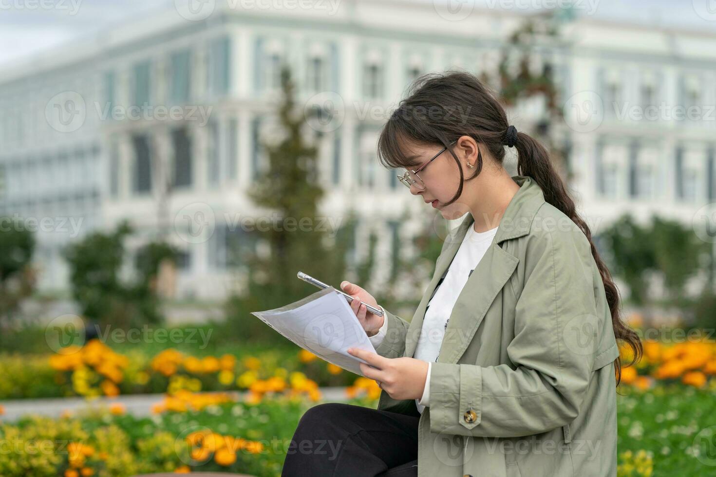 A girl sits on a bench on the street with documents in her hands and using her phone photo