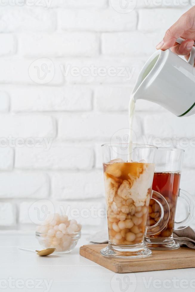 verter leche a mano en un vaso con té de burbujas helado, fondo de pared de ladrillo blanco foto