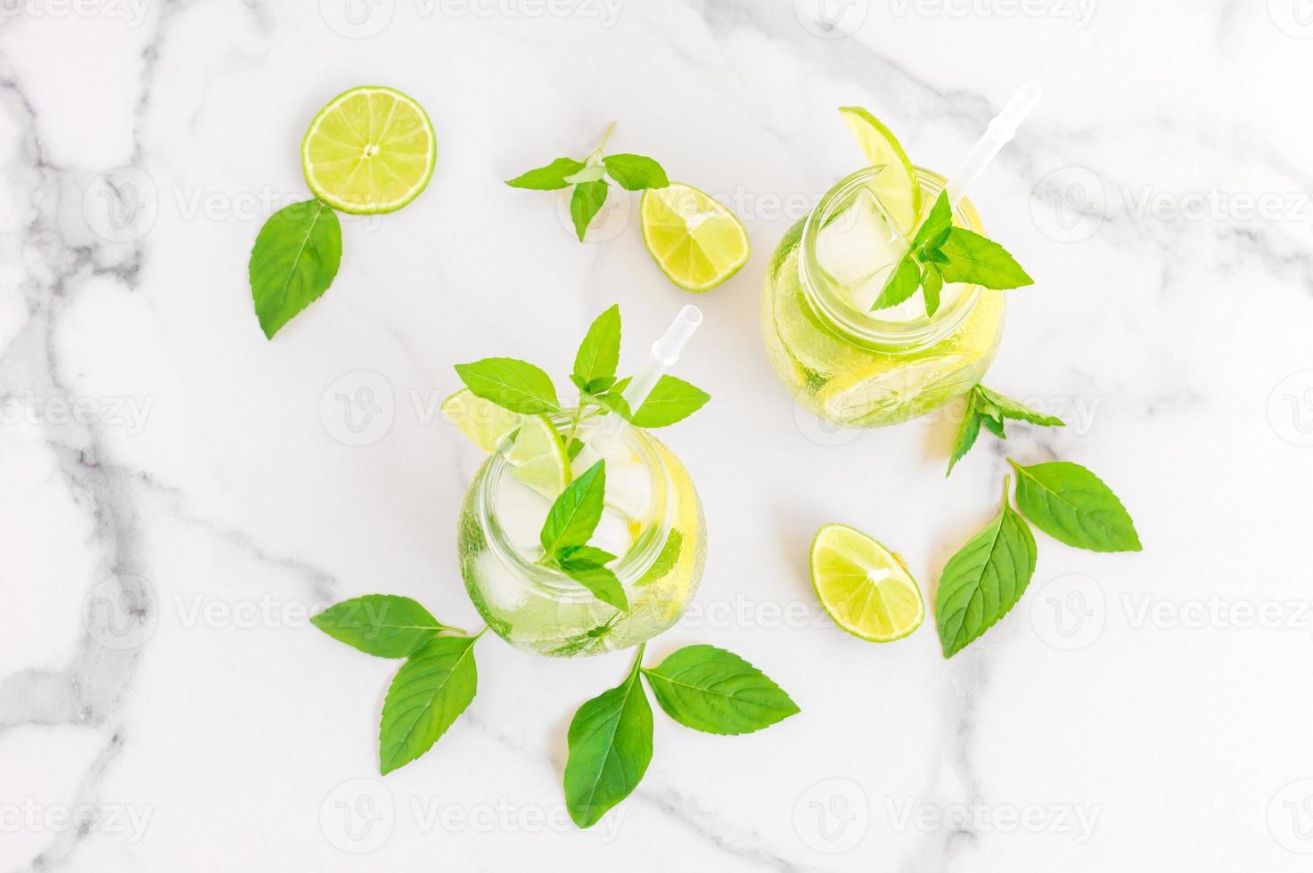 Two glasses with lemonade or mojito cocktail with lemon and mint, cold refreshing drink or beverage with ice on white marble background. photo
