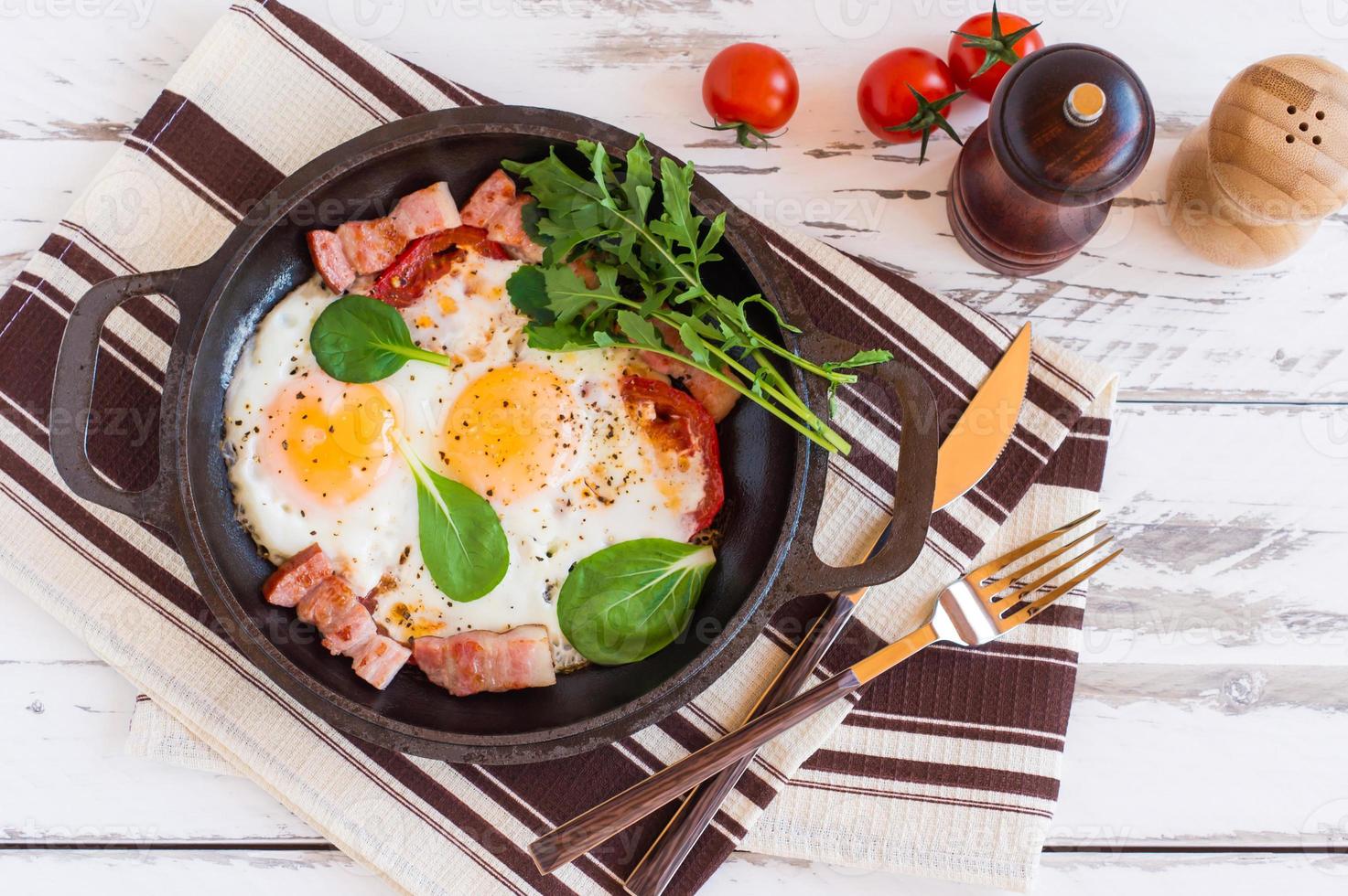 English breakfast or lunch with fried egg, spinach, arugula, tomatoes and bacon on black pan. photo