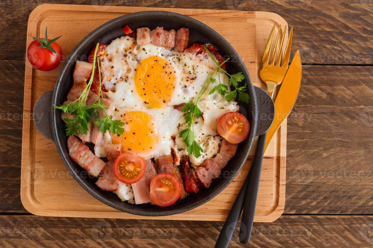 Homemade fried eggs with bacon and tomatoes in a frying pan on wooden background. Classic breakfast. Top view. photo