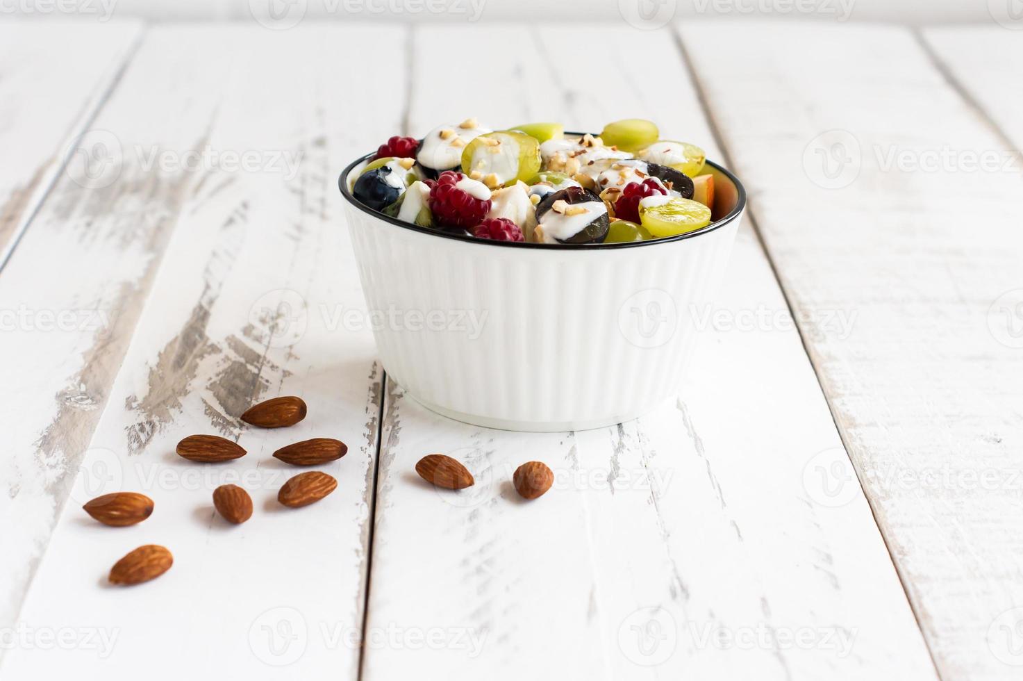 a bowl of fruit salad with yogurt and almonds on a white wooden table. diet breakfast. photo