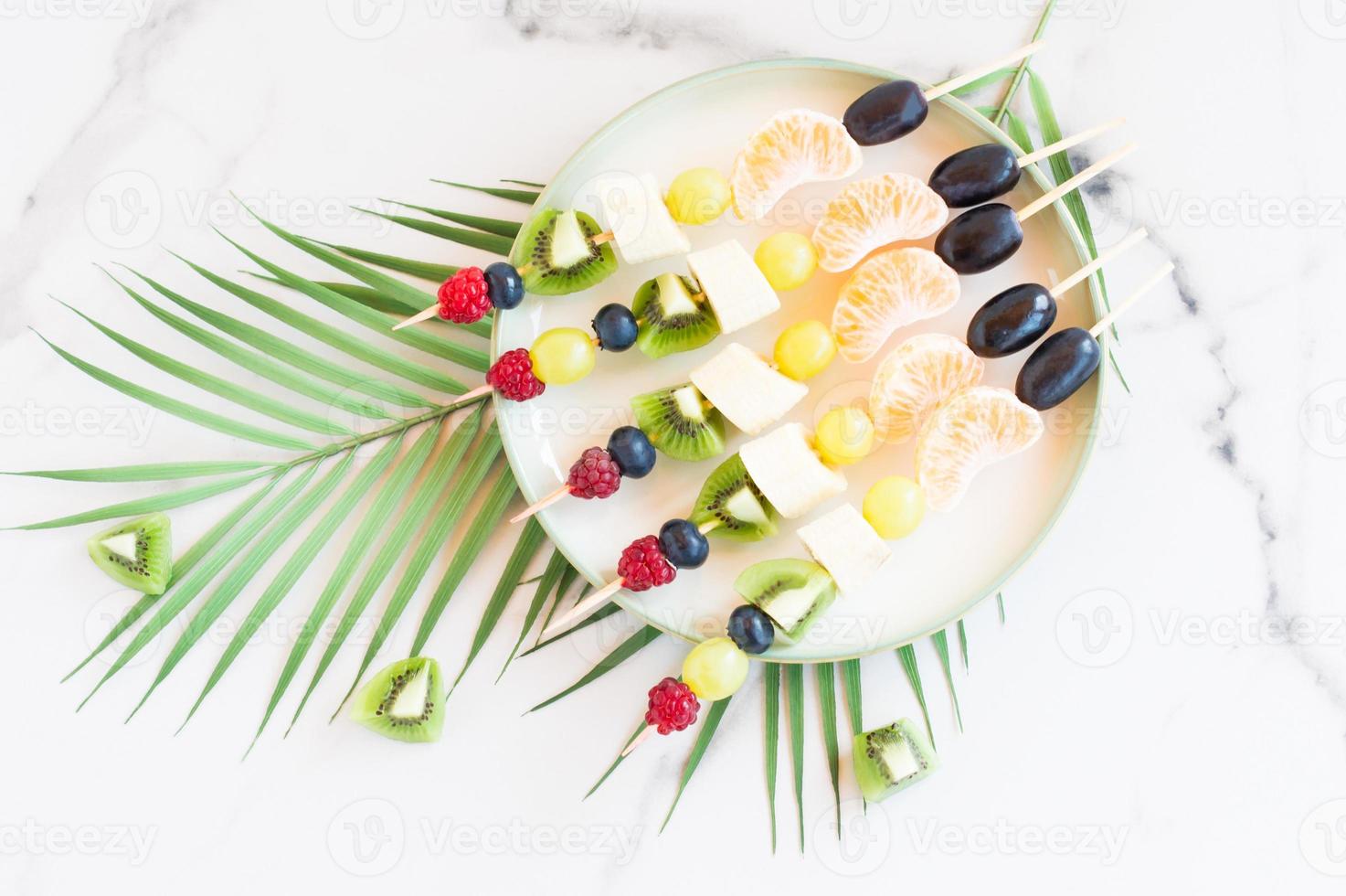 canapes of fresh fruit and berries on a plate. Marble background with palm tree branch. Festive food. photo