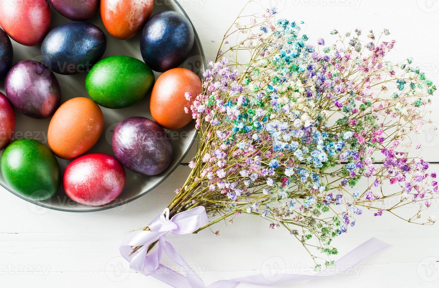 painted Easter eggs on a plate with a bouquet of bright flowers on a white wooden table. top view. photo