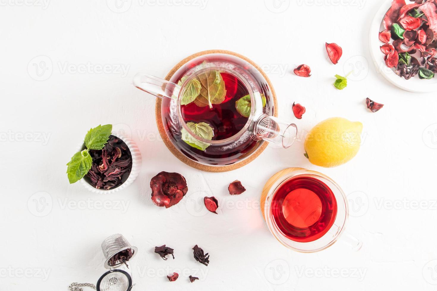 a teapot of freshly brewed hibiscus tea with mint leaves and lemon slices, a glass cup with hibiscus rose petal tea on a white background. photo