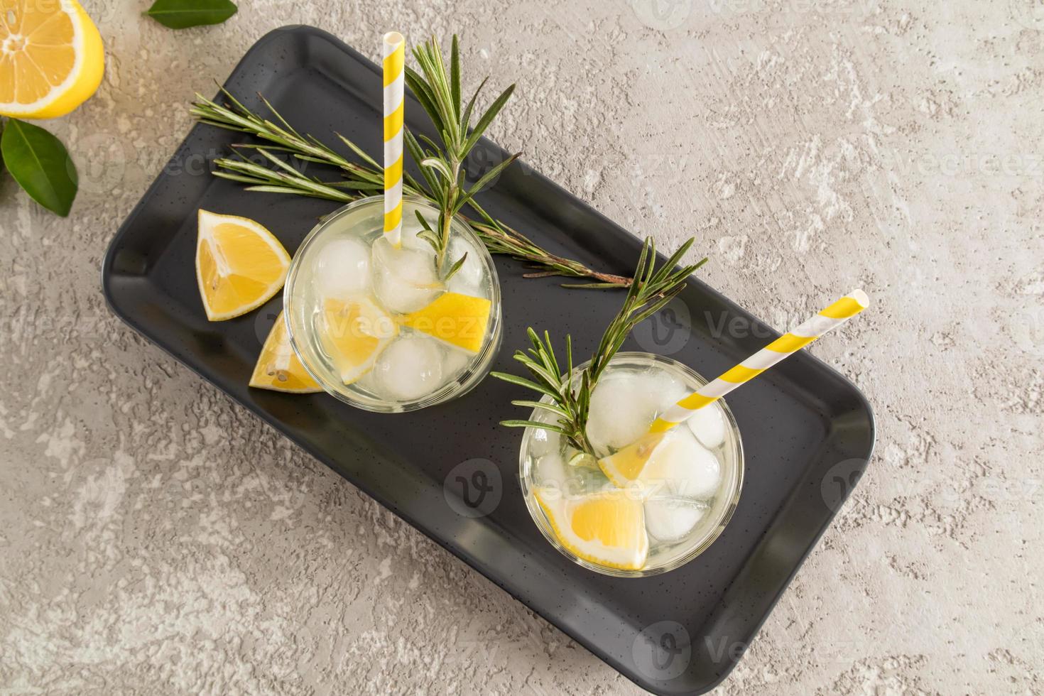 top view of two glasses of sparkling water with lemon and ice cubes on a ceramic tray and concrete table. fruit water is a concept of health. photo