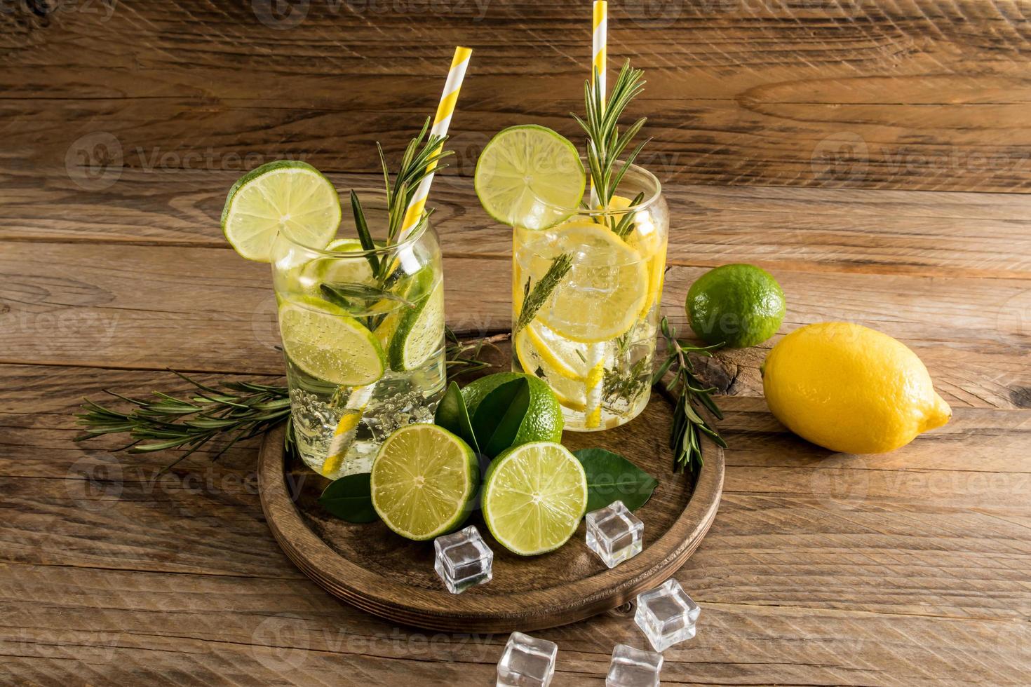 two glasses of vitamin water with lemon and lime, sprigs of rosemary on a round wooden tray and a village table. ice cubes for cooling. photo