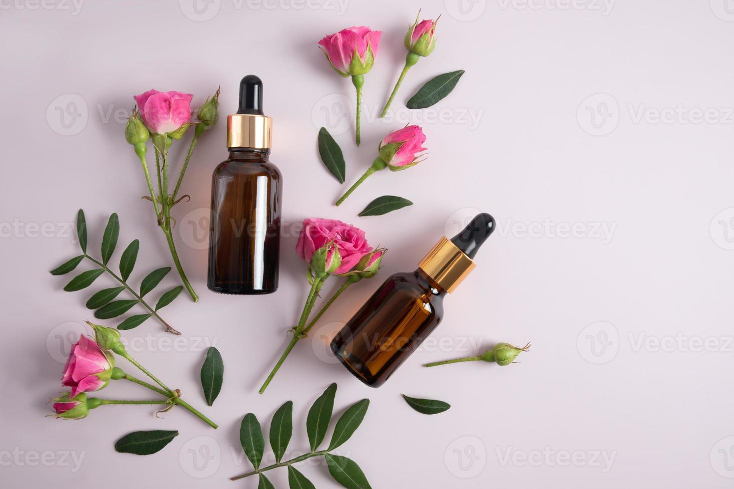 top view of two glass bottles with cosmetics, face serum against the background of pink rose buds and green leaves. photo