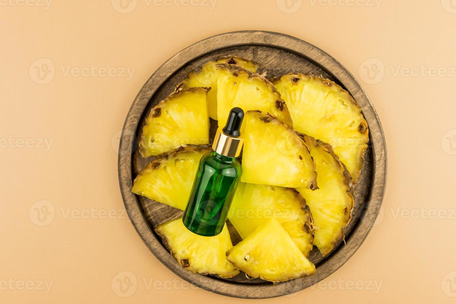 a wooden tray with slices of ripe pineapple and a cosmetic bottle with a green glass pipette. moisturizing cosmetics. top view. brown background. photo