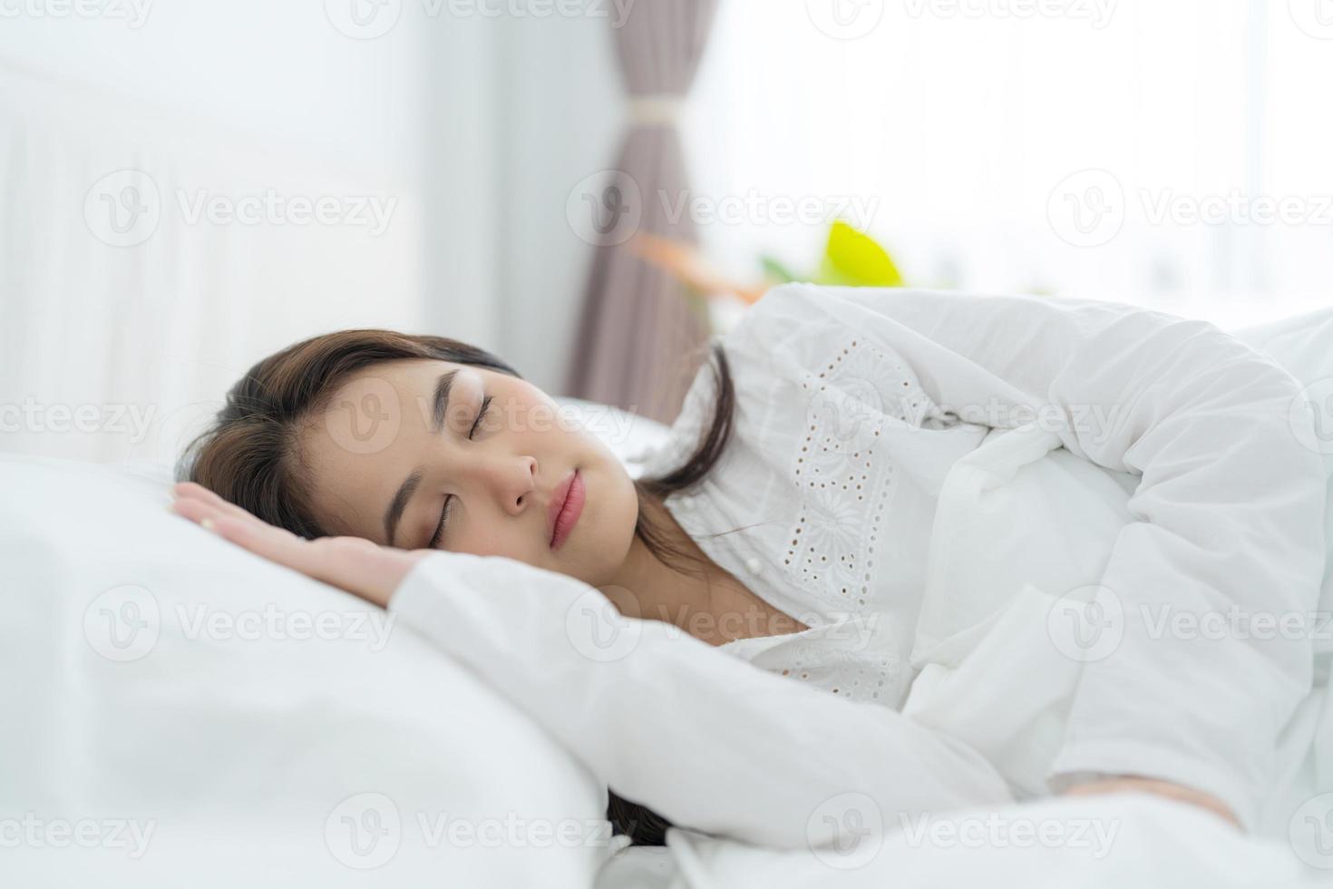 A beautiful woman lays her hands on a soft pillow. A young woman wearing white pajamas lies under a warm blanket. photo