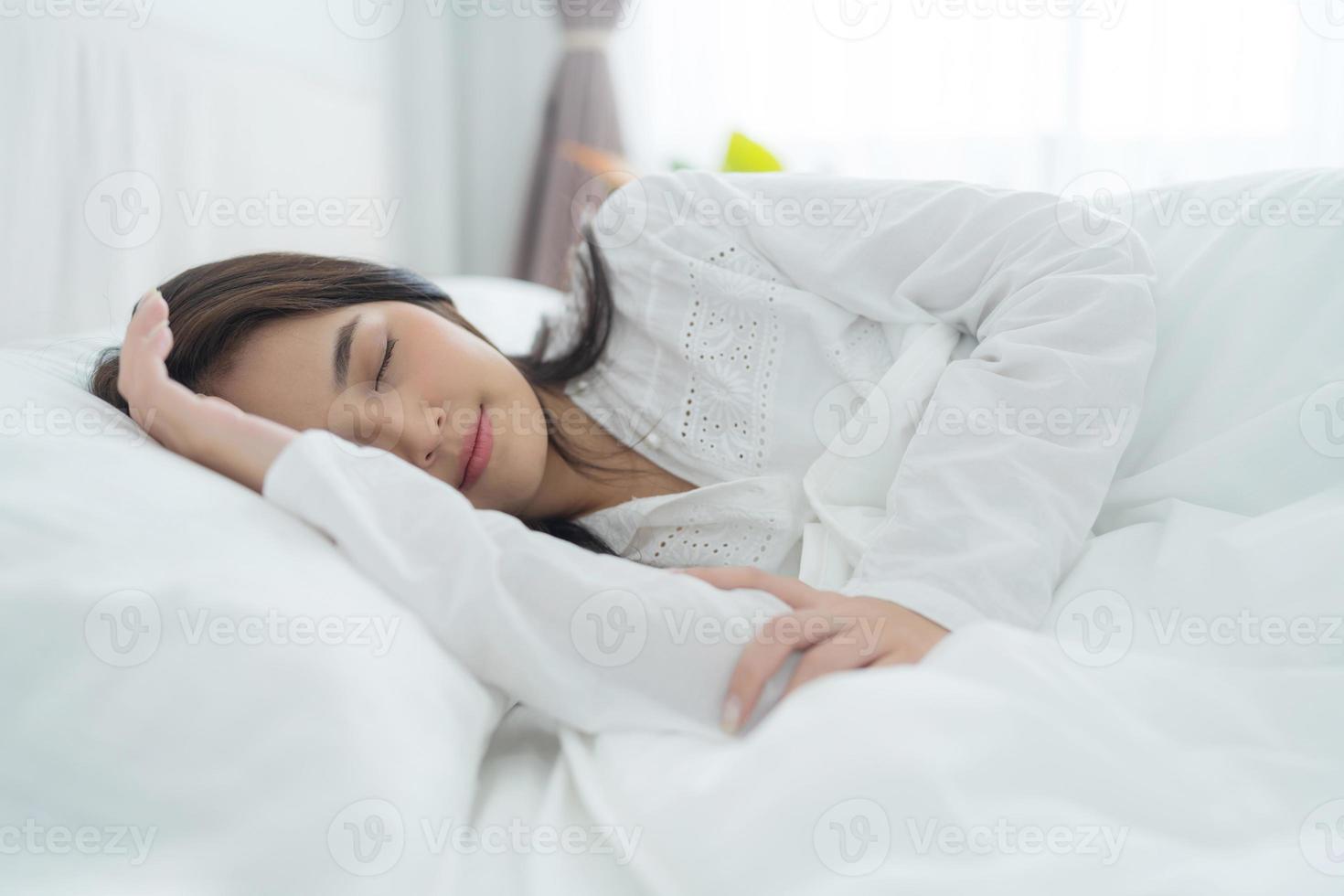 A beautiful woman lays her hands on a soft pillow. A young woman wearing white pajamas lies under a warm blanket. photo