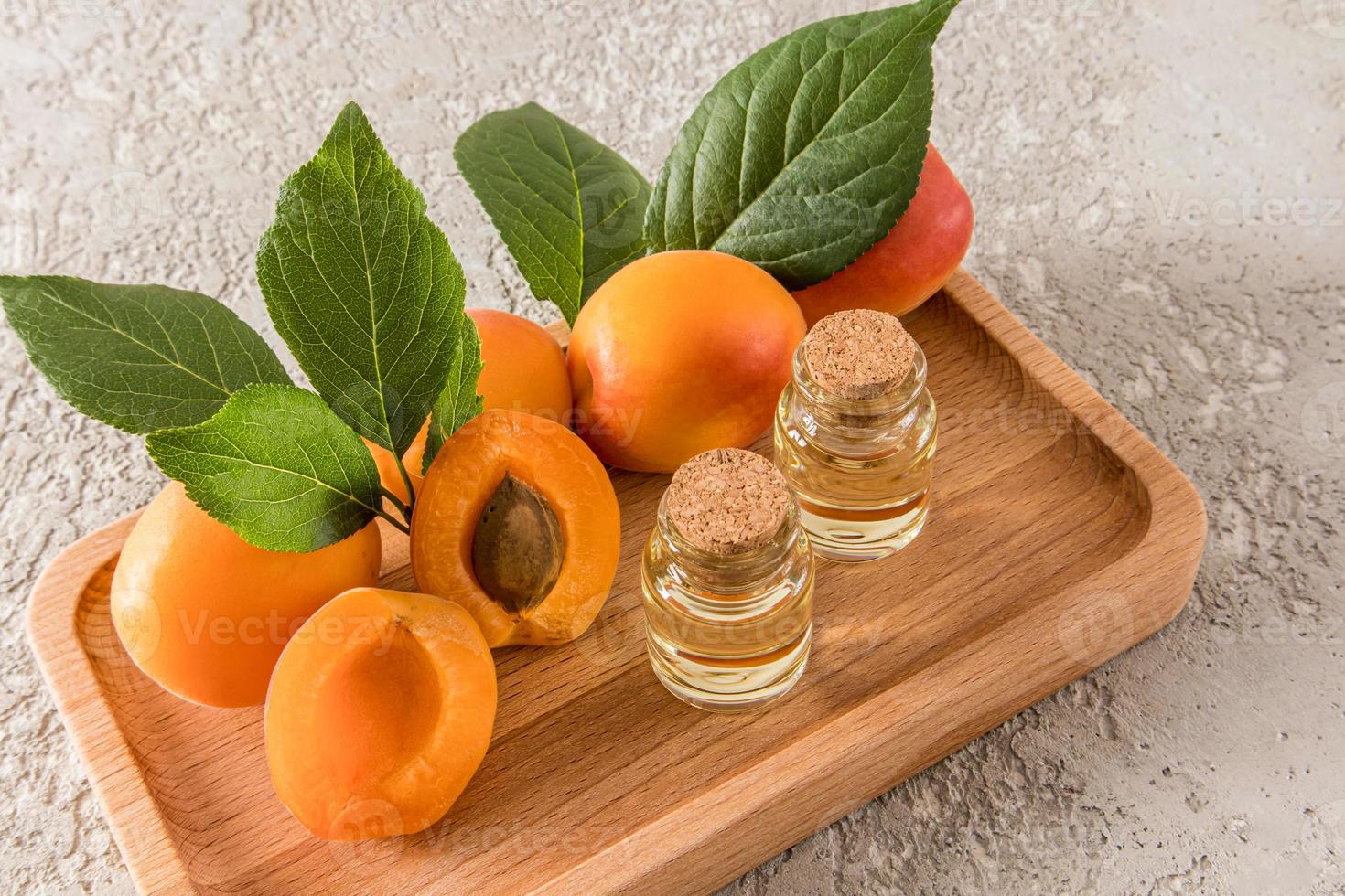 two glass bottles with cork cap with apricot kernel oil among fruits and juicy apricot leaves on the tray. gray cement background. photo