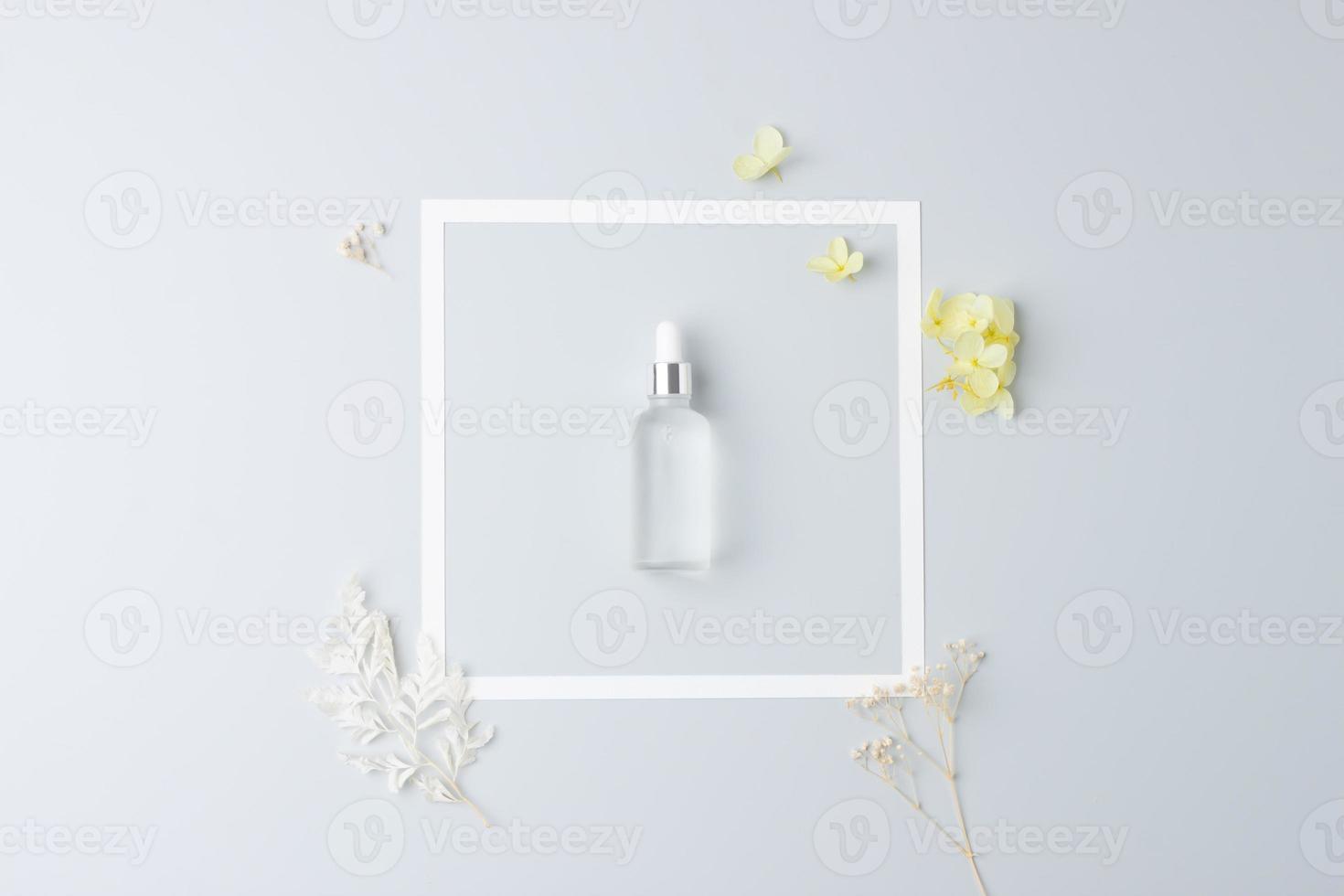 Cosmetic bottle with flowers and white frame on grey background. Flat lay, copy space photo