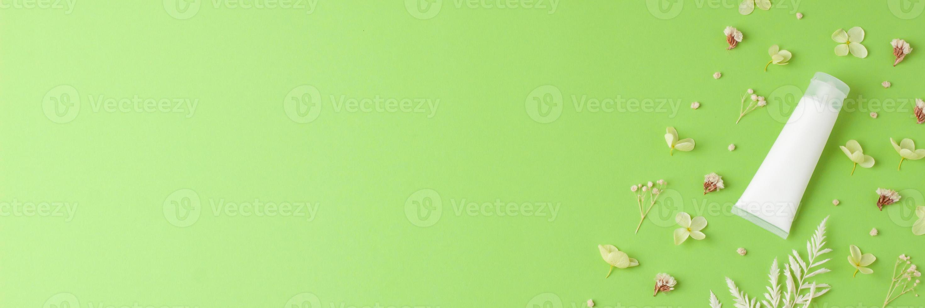 White tube of cosmetic cream with flowers on green background. Flat lay, copy space photo