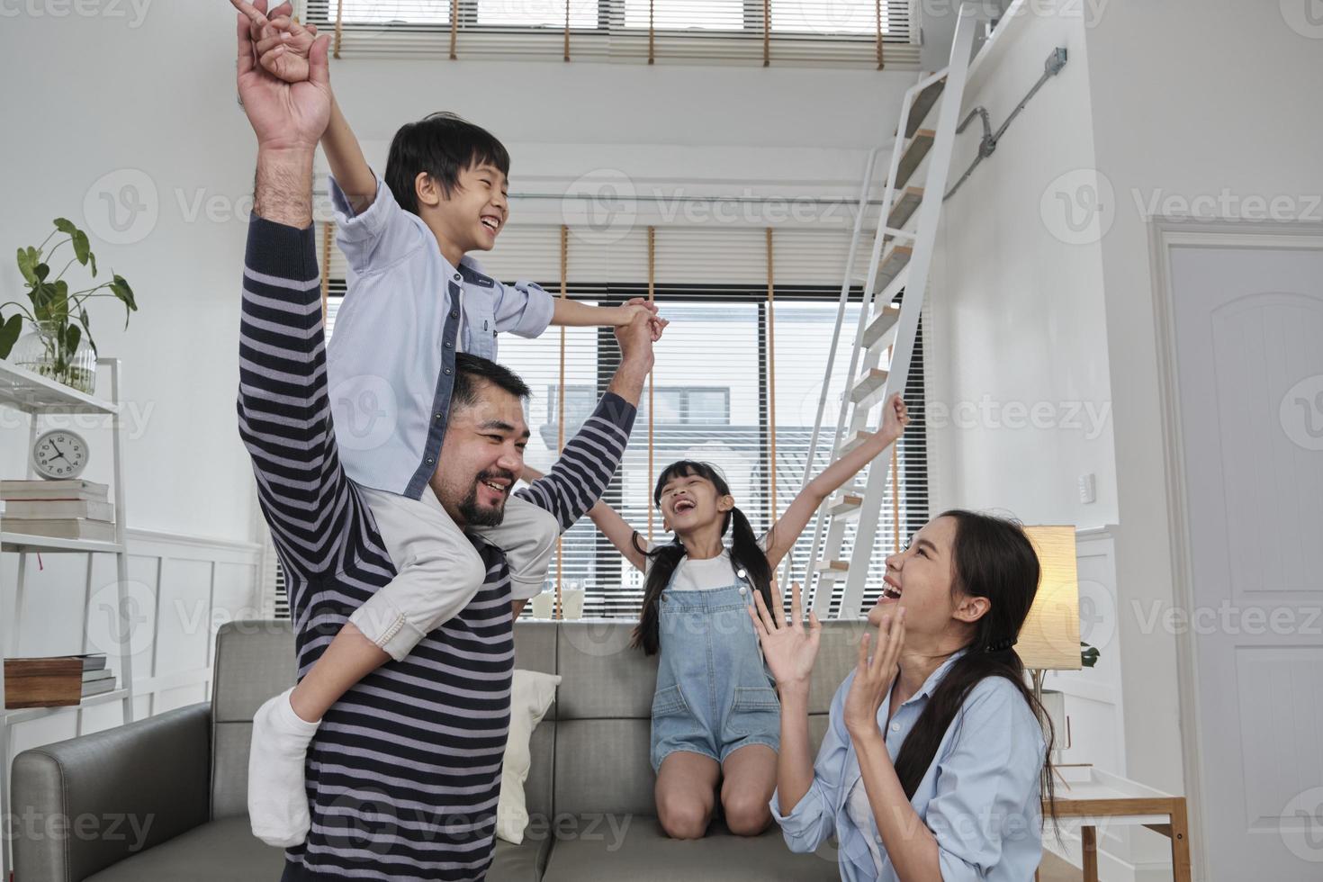 Asian Thai family together, dad plays and teases with children and mum by carrying and holding son on shoulders in home living room, happy leisure times, lovely weekend, wellbeing domestic lifestyle. photo