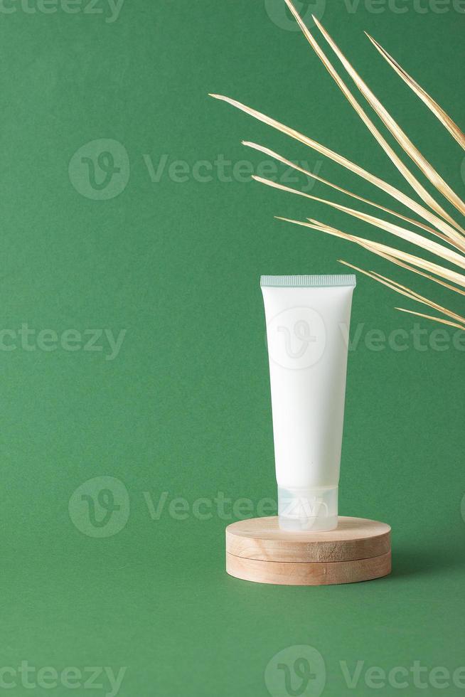 White tube of cosmetic cream with wood podium and palm leaf on green background. Close up, copy space photo