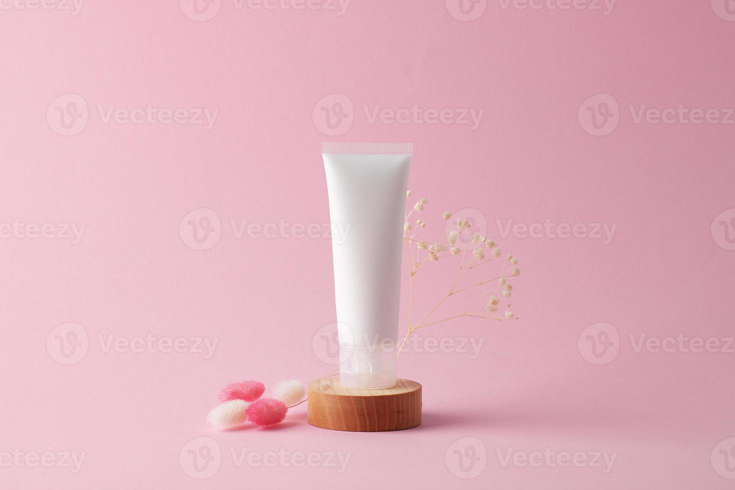 White tube of cosmetic cream with flowers on wooden podium on pink background. Close up. photo