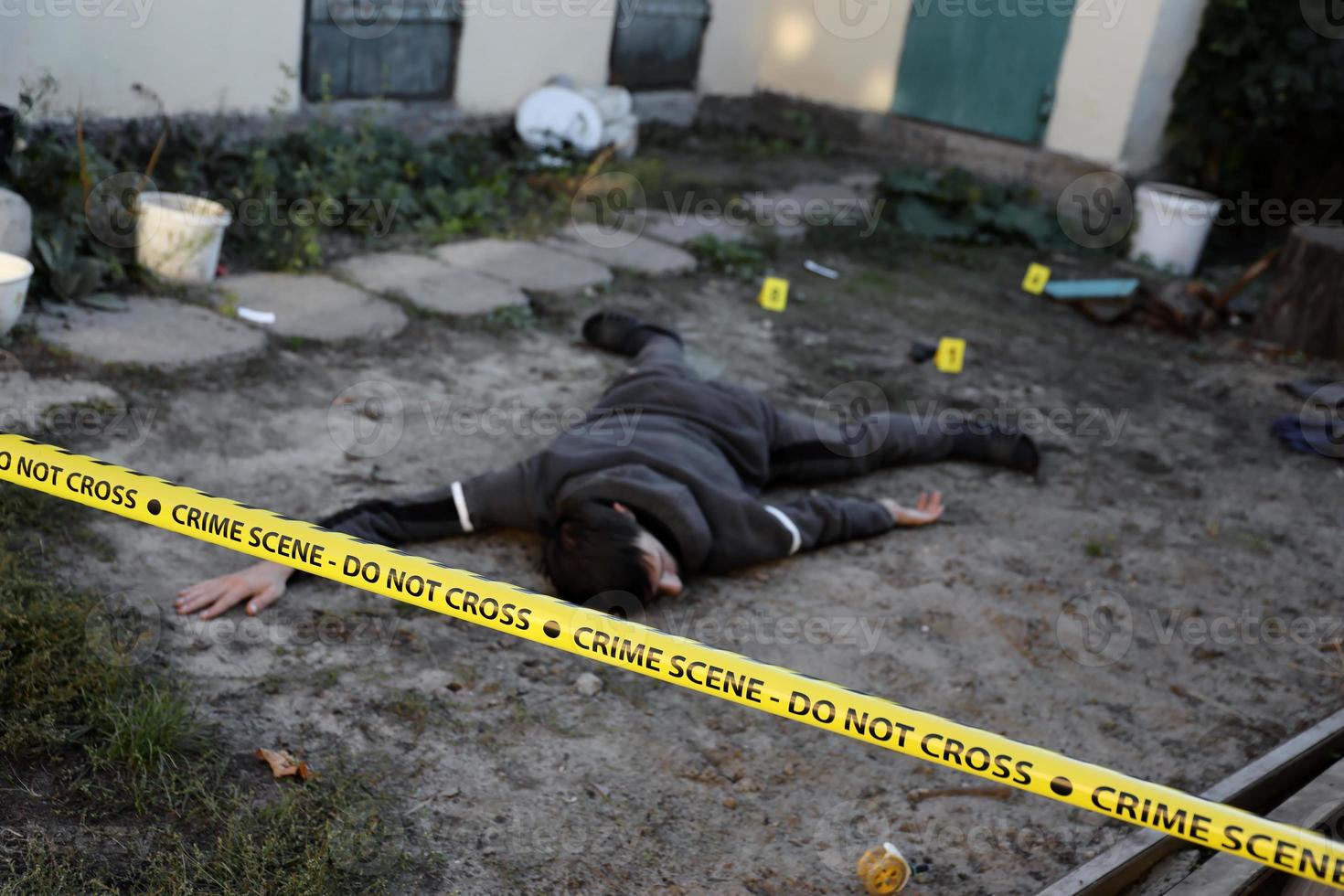 Victim of a violent crime in a backyard of residental house in evening. Dead man body under the yellow police line tape and evidence markers on crime scene photo