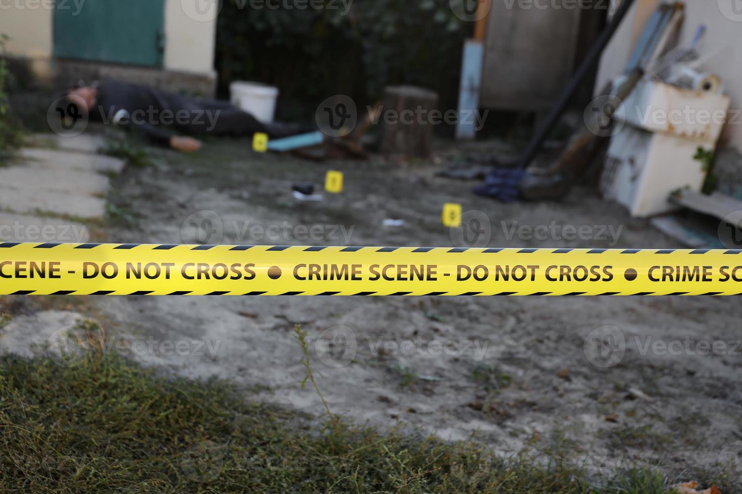 Victim of a violent crime in a backyard of residental house in evening. Dead man body under the yellow police line tape and evidence markers on crime scene photo