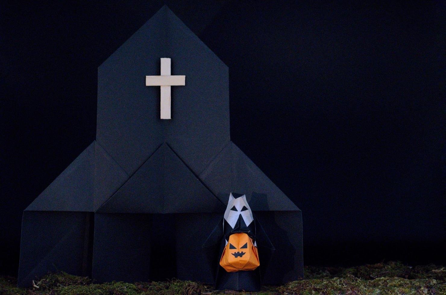 The Halloween background of origami or Paper folding about the nun holding pumpkin head jack-o-lantern in front of black church with messy lawn photo