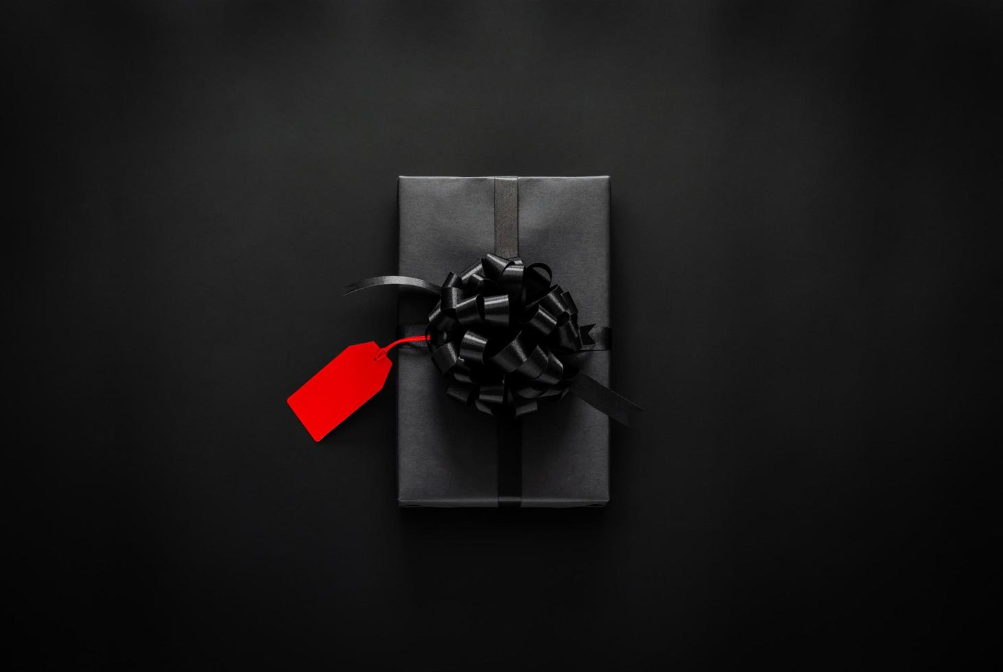 A black gift box with red ribbon and blank price tag puts on black background. Black friday and Boxing day concept. photo