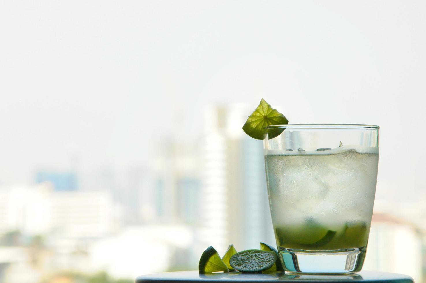 un vaso de caipirinha con la rodaja de lima mezclada con jugo de lima, azúcar, cachaca o ron brasileño y cubitos de hielo para disfrutar del clima cálido en verano con el fondo de la vista de la ciudad. foto