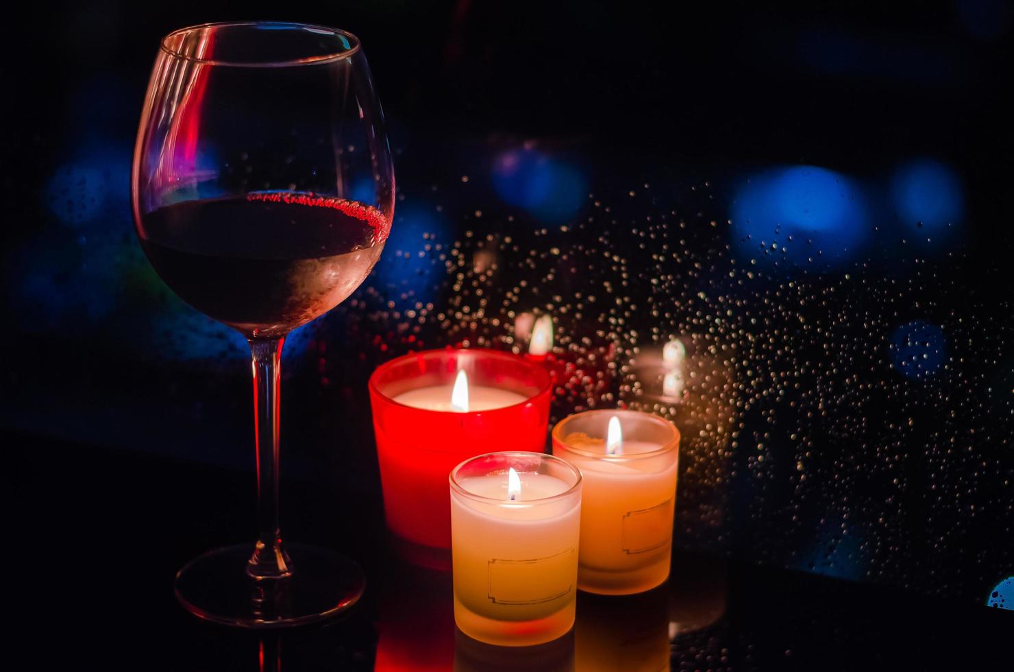 A glass of red wine with burning candles put beside window that have rain drop on dark background. photo